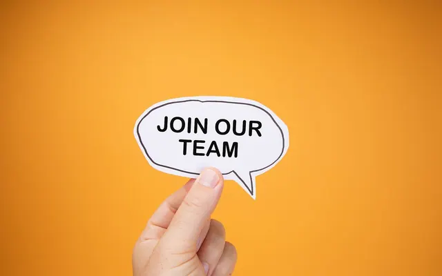 Human Hand Raises Paper Speech Bubble with JOIN OUR TEAM text Against Orange Backdrop (53181686992)