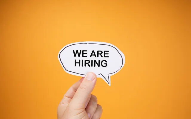 Human Hand Raises Paper Speech Bubble with WE ARE HIRING text Against Orange Backdrop (53182280041)
